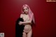 A naked woman with pink hair posing in front of a red wall.